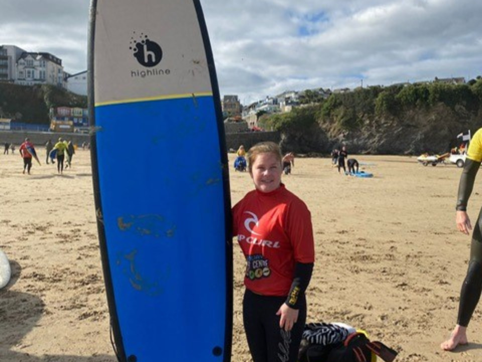 Sassy is standing on a beach holding a blue surfboard and smiling. She’s wearing a wetsuit and a red rash guard that says rip curl on it. Behind her you can see cliffs and other people on the beach