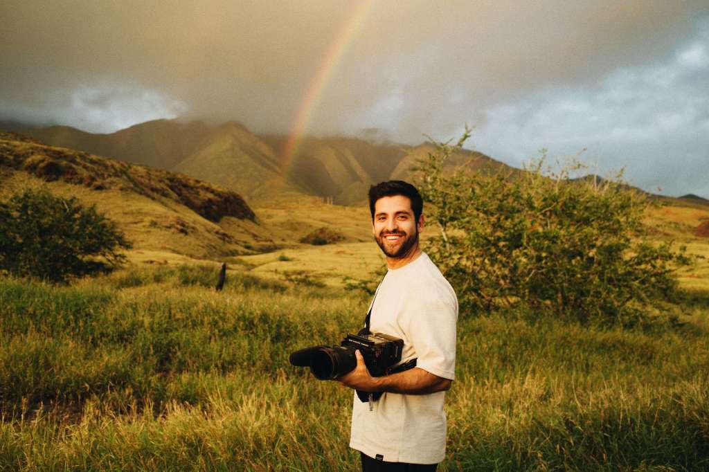 Matias de Rada holding a camera in Hawaii with a rainbow in the background.