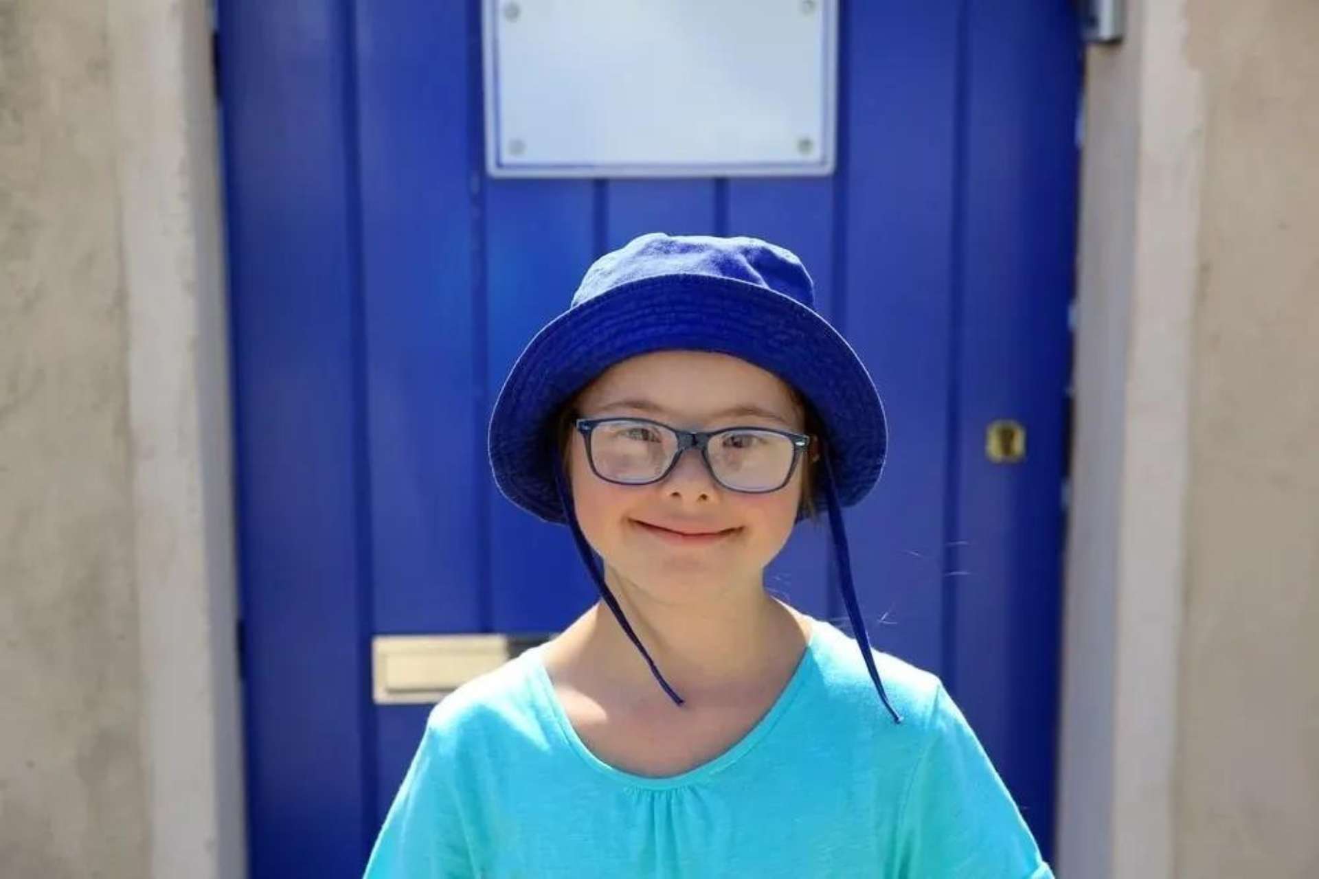 Girl smiling at the camera wearing a sun hat. She is standing on front of a blue door