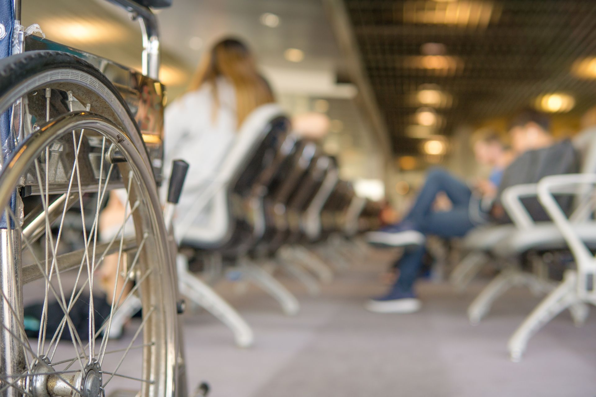 In the foreground of this airport departure area is a service wheelchair. In the background several defocussed people are waiting in the seating area for a flight.